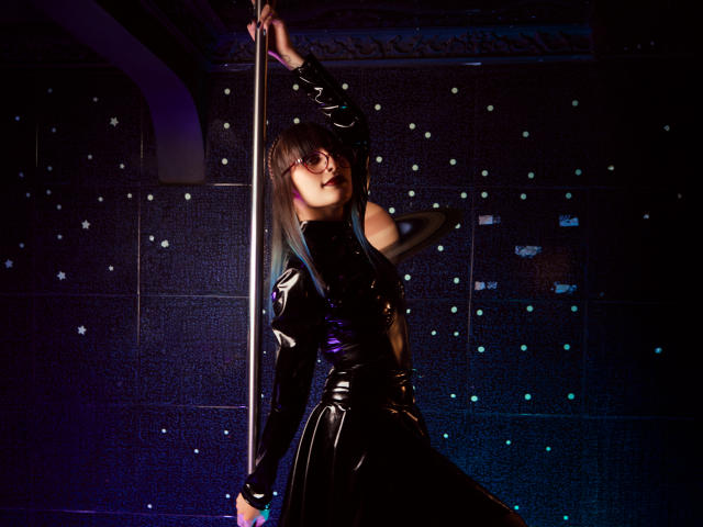 TatsumiLee, dressed in tight, shiny black latex, grips a pole with one hand while leaning back, legs slightly apart. Dark, glittery background, intense lighting, provocative pose, sexy.