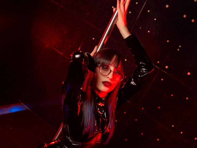 Digitale Kunst von TatsumiLee, mit langen, schwarzen Haaren, roten Strähnen und rotem Lippenstift, die ein glänzendes, schwarzes Latex-Outfit trägt. Outfit, der eine silberne Stange hält, im Hintergrund leuchten rote Lichter. Verführerischer, intensiver Ausdruck.