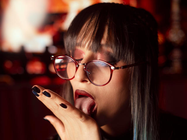Close-up photo of TatsumiLee with black hair, red glasses, and black nail polish. TatsumiLee's tongue is out, licking their index finger seductively. Background is blurred with red and orange lights.