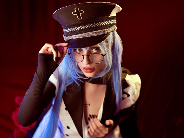 TatsumiLee, dressed as a sexy nurse, adjusts a black and white military-style cap with a cross. Blue hair, glasses, black and white outfit, red lips, intense gaze, dark background.