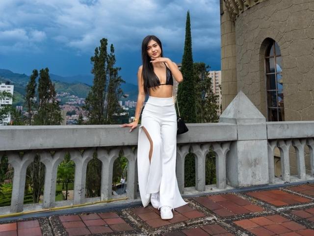 MiaaFerrer, attraente, in piedi su un balcone, pantaloni bianchi, top nero con spalline, lunghi capelli neri, appoggiata alla ringhiera, blu. sfondo del cielo, montagne, alberi, edificio in pietra, posa sicura, incredibilmente attraente..