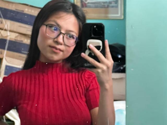 AnittaRouses, wearing a red ribbed turtleneck, takes a mirror selfie with a smartphone. She has long black hair, glasses, and a slight smile. Background includes wooden slats and a teal wall.