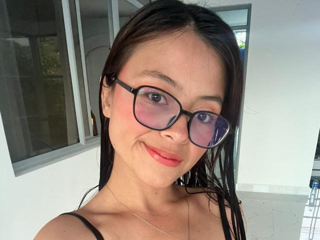 Close-up of AnittaRouses with long black hair, wearing black-framed glasses and a black spaghetti strap top. She's smiling with red lipstick, and there's a visible window in the background.