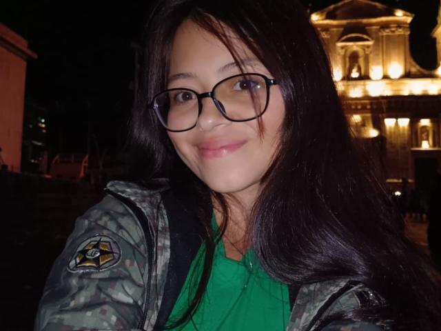 Close-up of AnittaRouses with long black hair, wearing black-rimmed glasses and a green shirt under a camo jacket. Background features brightly lit, ornate building. She smiles subtly.