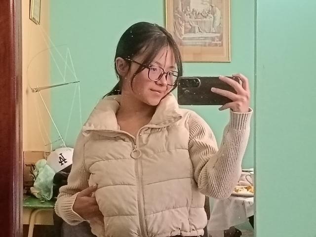AnittaRouses, an Asian woman with glasses, takes a mirror selfie. She's wearing a beige puffy vest, holding her belly, with a teal wall and framed picture in the background. Fucking cute.