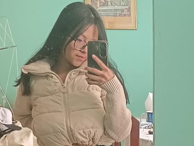 AnittaRouses, avec ses longs cheveux noirs et ses lunettes, prend un selfie devant un miroir dans une pièce bleu turquoise. Elle porte une veste matelassée beige et tient un téléphone noir. Elle est incroyablement attirante, avec un léger froncement de sourcils..