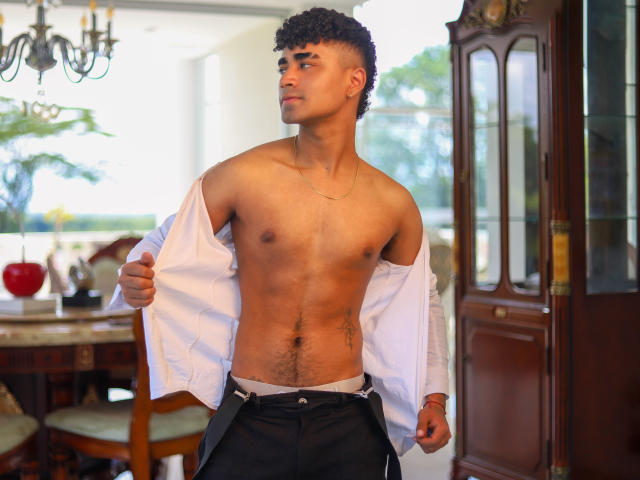 Jacobb69, a young man with curly black hair, stands shirtless in a room, pulling open a white shirt to reveal a toned, hairy chest. Wearing black pants, looking to the side.