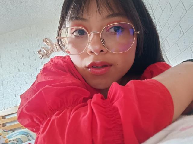 Close-up of AnittaRouses with straight black hair, wearing round glasses and a red puffy shirt, lying on a bed, mouth slightly open, soft light on white tiled wall. Fucking cute.