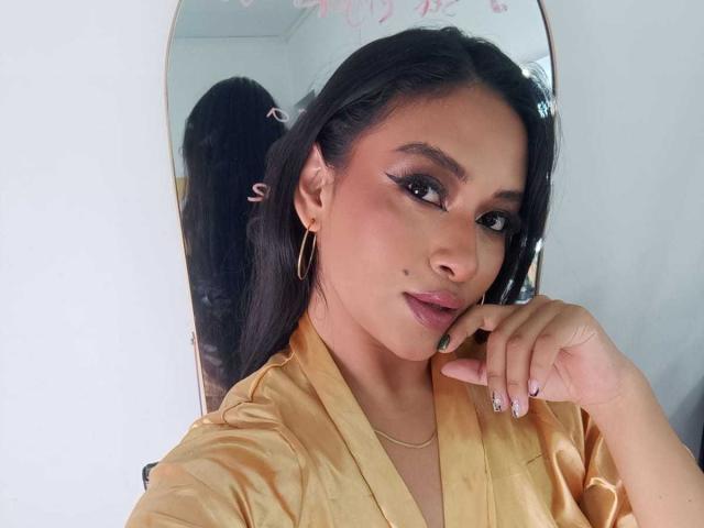 Close-up selfie of AdalinaVera, an attractive woman with long black hair in a ponytail, wearing a yellow satin top, large hoop earrings, and dark lipstick. She touches her chin suggestively.