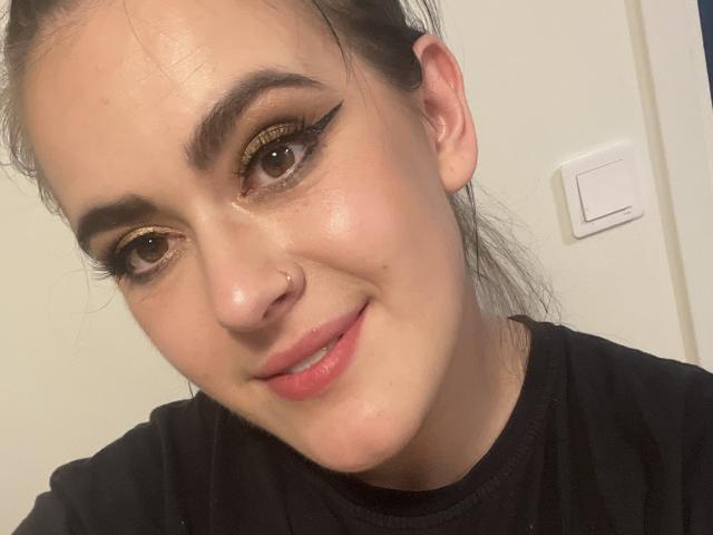 Selfie de primer plano de Diora, una mujer blanca con cejas oscuras, sombra de ojos dorada y delineador de ojos negro. Ella tiene un pequeño piercing en la nariz, cabello oscuro y lleva una camisa negra. Muy atractivo..