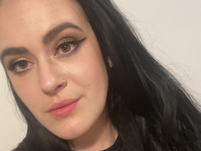 Close-up van Diora, een jonge vrouw met bleke huid, zwart haar en bruine ogen. Ze heeft een neusring, opvallende eyeliner, roze lippen en een subtiele grijns. Heel aantrekkelijke uitstraling.