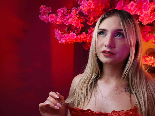 Fotografia di MariyLu con lunghi capelli biondi, una camicetta rossa in pizzo e un piccolo tatuaggio sul petto, che tiene in mano un fiore rosso. Uno sfondo vivace con fiori di ciliegio rossi e rosa. Molto attraente..