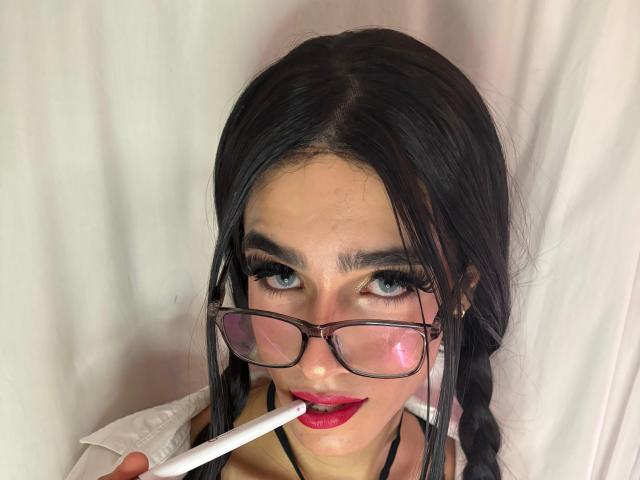 Fotografía de primer plano de SophieLondonQr, una mujer con cabello negro largo trenzado, que usa gafas, y está chupando un palito blanco, rojo. pintalabios, y maquillaje intenso, fondo blanco, tensión sexual.