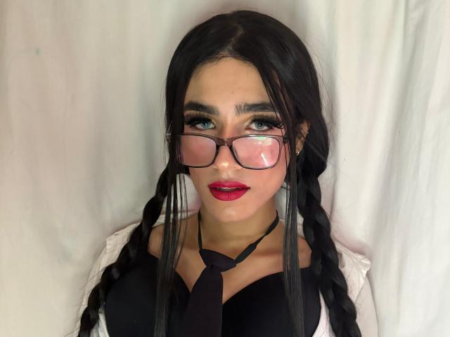 Fotografie von SophieLondonQr, einer attraktiven Frau mit langen schwarzen Zöpfen, rotem Lippenstift und schwarzen Brillen. Sie trägt ein schwarzes Oberteil, eine weiße Jacke und eine schwarze Krawatte und strahlt eine sexuelle Spannung aus..
