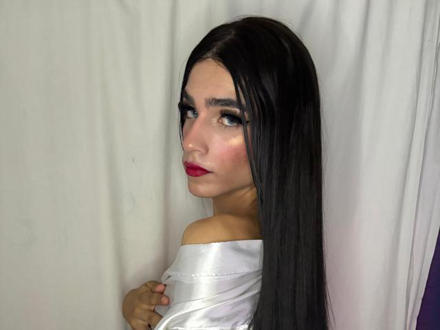 Foto van SophieLondonQr, met lang zwart haar, rode lippenstift en een witte top met een open schouder, die om zich heen kijkt met een verleidelijke blik. uitdrukking.
witte achtergrond met een gordijn. Sensueel, verleidelijk, intense blik..