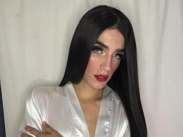 Fotografie von SophieLondonQr mit langen, schwarzen Haaren, die ein glänzendes, weißes Oberteil, roten Lippenstift und intensive blaue Augen trägt, Arme durchgestrichen, Hintergrund weiß. Die sexuelle Spannung ist in ihrem direkten Blick deutlich erkennbar..