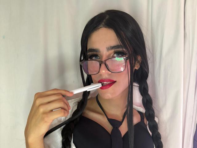 Fotografia di SophieLondonQr, una giovane donna con lunghe trecce nere, che indossa occhiali, una maglietta nera e lecca qualcosa di bianco. gelato alla frutta. Le sue labbra rosse sono leggermente dischiuse, creando un'espressione seducente. Illuminazione intensa.