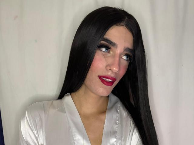Foto van SophieLondonQr, een aantrekkelijke vrouw met lang zwart haar, die een glanzend wit blouse draagt en naar de zijkant kijkt. een lichte glimlach, rode lippen en een verleidelijke blik.