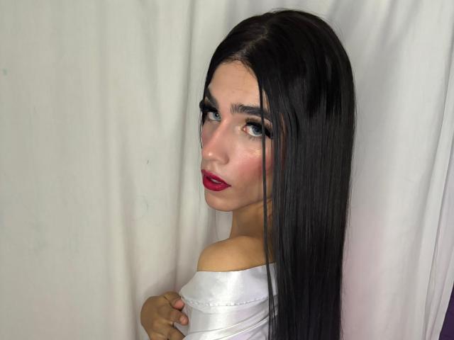 Photograph of SophieLondonQr, a sexy woman with long black hair, red lipstick, and white off-shoulder top, looking back with a sultry expression. Her hand touches her chest, showing tension.