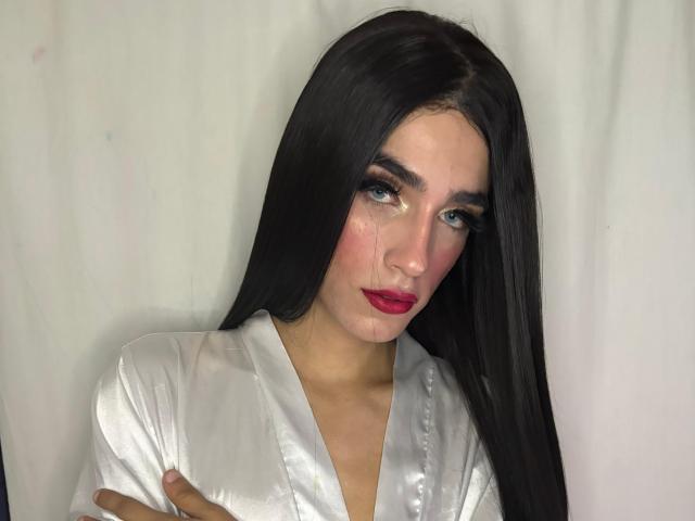 Foto van SophieLondonQr, een aantrekkelijke vrouw met lang zwart haar, die een glanzend wit top draagt en recht in de camera kijkt. camera met rode lippenstift en uitgesmeerde make-up. Extreem intense blik.
