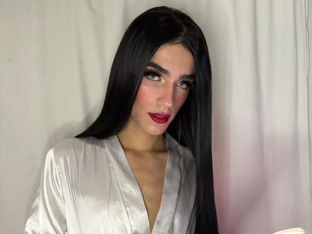 Fotografia de SophieLondonQr, uma mulher atraente com longos cabelos pretos, vestindo uma blusa branca brilhante com decote em V, batom vermelho e maquiagem para olhos com efeito esfumado. Ela olha de forma sedutora, com uma tensão sexual evidente..