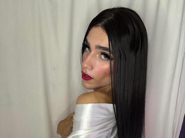 Foto van SophieLondonQr, een aantrekkelijke vrouw met lang zwart haar, rode lippenstift en donkere ogen, die over haar schouder kijkt. Ze is gehuld in een glanzende witte doek, waardoor haar schouders zichtbaar zijn..