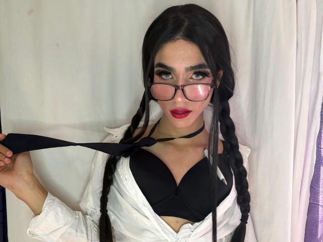 Fotografía de SophieLondonQr, una mujer de pelo oscuro con trenzas en forma de coletas, que lleva gafas, una camisa blanca y un sujetador negro, sosteniendo traje formal negro, labios rojos, fondo de cortina blanca, mirada intensa, tensión sexual, pose seductora..