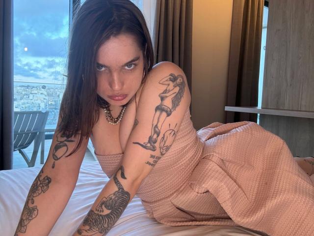 ZoeFox-VIP-hot, femme très tatouée avec de longs cheveux bruns, un regard intense, se penchant de manière séduisante sur un lit, vêtue d'une robe couleur pêche. Pièce lumineuse, vue sur la ville à travers la fenêtre, pose sexy et provocante, lumière naturelle..