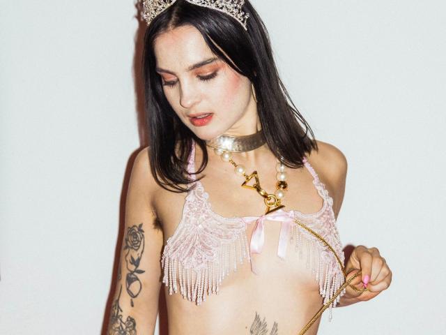 ZoeFox-VIP-hot, com longos cabelos pretos, vestindo um top rosa com franjas, uma tiara com orelhas de gato e um colar dourado. Ela tem tatuagens no braço esquerdo e na parte inferior do abdômen, olhando para baixo de forma sedutora..