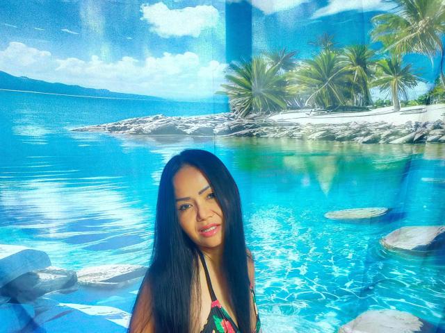 Lotusblume, de piel morena y con el pelo largo y negro, vestida con un bikini colorido, está de pie en un agua azul brillante con palmeras en el fondo. Su expresión es ligeramente seductora, con una luz solar vibrante. Hermosamente increíble..