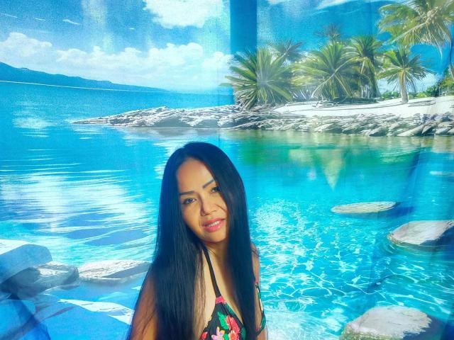 Lotusblume, une jeune femme aux longs cheveux noirs, vêtue d'un bikini coloré, se tient dans une eau bleu vif et sourit. Des palmiers et des rochers en arrière-plan, une lumière bleue et blanche intense, une ambiance tropicale..