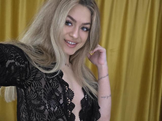 Fotografía de NayaQuinn, una mujer rubia con cabello largo, que lleva una blusa negra de encaje con un escote pronunciado y está sonriendo directamente a la cámara. enfrentado a la cámara, cortinas amarillas en el fondo, mano cerca del rostro.