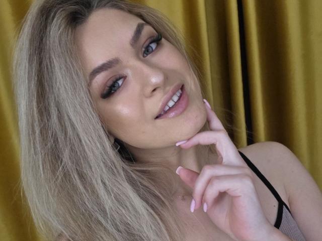Close-up de NayaQuinn, uma mulher loira com olhos azuis, lábios cheios e unhas rosadas, tocando o queixo com a expressão sensual, vestindo uma blusa preta com alças, cortinas douradas ao fundo..