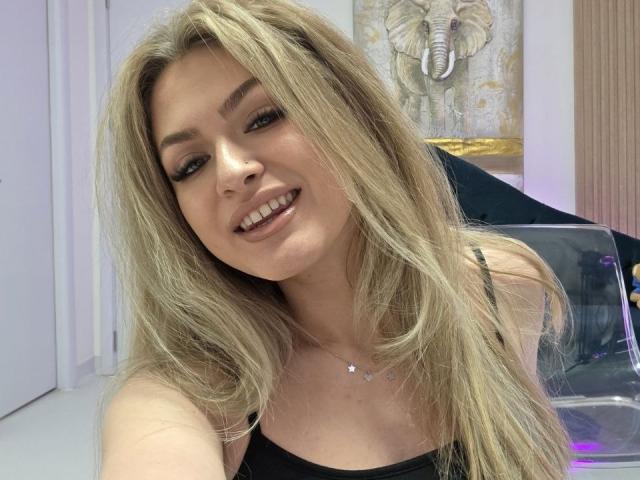 Nahaufnahme von NayaQuinn, blonde Haare, lächelnd, schwarzes Tanktop, sichtbarer Ausschnitt, helle Haut, dezentes Make-up, Hintergrund enthält Ventilator und Wand, sexuelle Spannung durch direkten Blick und leicht geöffneten Mund..