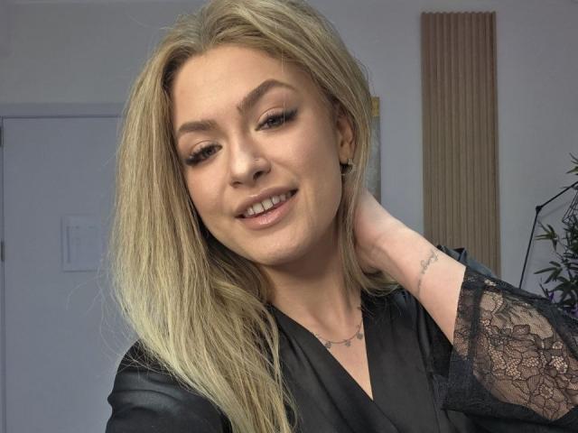 Fotografia di NayaQuinn, una donna bionda con lunghi capelli, che indossa una camicetta nera in pizzo, sorridente con gli occhi socchiusi, a destra. mano dietro la testa, illuminazione soffusa, leggera tensione sessuale, posa informale.