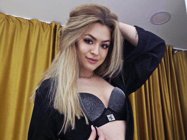 NayaQuinn, une femme blonde aux longs cheveux, porte un soutien-gorge en dentelle noire et un cardigan noir, une main dans les cheveux, avec des rideaux jaunes en arrière-plan. fond, expression sensuelle, lèvres légèrement entrouvertes, pose suggestive.