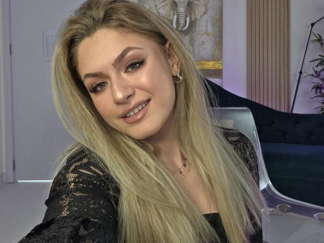 Foto van NayaQuinn, een blonde vrouw met lang haar, een zwarte kanten top, die lichtjes glimlacht, bruine ogen, subtiele make-up, neus. ring, oorbellen, zacht licht, modern interieur, subtiele seksuele spanning, nonchalante pose.