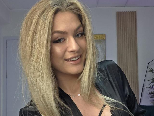 Close-up of NayaQuinn, a blonde woman with straight hair, smiling slightly, wearing a black top, showing cleavage, in a well-lit room. She has light skin, blue eyes, and a confident look.