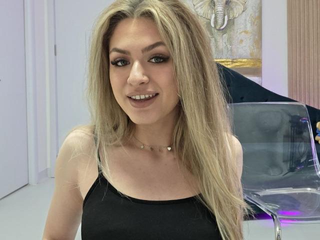 Nahaufnahme von NayaQuinn, einer blonden Frau mit langen Haaren, die ein schwarzes Tanktop trägt und lächelt. Sie hat dunklen Eyeliner und eine Nase. Piercing. Ihre Halskette hat kleine, herzförmige Anhänger. Im Hintergrund ist ein Stuhl zu sehen..