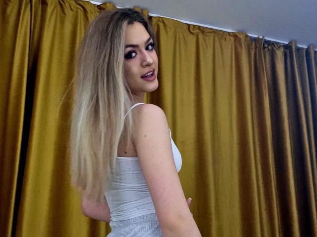 NayaQuinn, rubia con cabello largo, vestida con una camiseta blanca ajustada, mirando por encima del hombro con una sonrisa seductora. Cortinas doradas en el fondo, una luz tenue proveniente de arriba. Increíblemente atractivo/a..