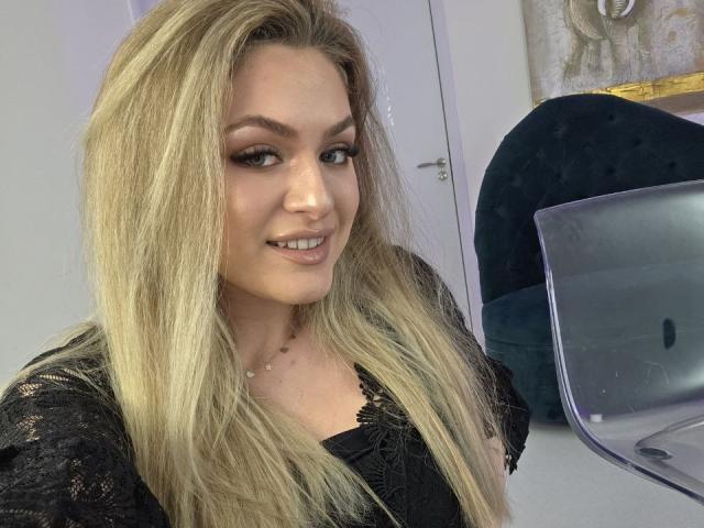 Fotografie von NayaQuinn, einer blonden Frau mit langen Haaren, die ein schwarzes Oberteil trägt und verführerisch in die Kamera lächelt. Ein Laptop und ein schwarzer Stuhl im Hintergrund. Verdammt heiß.