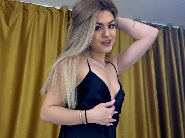 NayaQuinn, avec ses longs cheveux blonds, porte une robe noire à décolleté plongeant, qui révèle sa poitrine. Elle pose, une main derrière la tête et l'autre sur sa poitrine, avec des rideaux jaunes en arrière-plan. Extrêmement sexy..