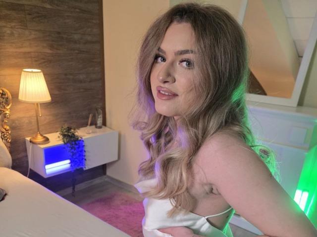 Naya Quinn, com longos cabelos castanhos ondulados, está sentada em uma cama, vestindo uma blusa branca sem alças, olhando por cima do ombro. Expressão sensual. A luz quente de um abajur a ilumina. Uma luz neon verde brilha ao fundo..