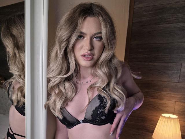 Fotografía de NayaQuinn con cabello rubio largo y ondulado, vistiendo un sujetador de encaje negro, de pie frente a un espejo, con su reflejo. luz, y una lámpara con luz cálida sobre una pared de madera. Tensión sexual.