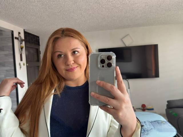 Lauren Spitz, een blanke vrouw met lang, steil bruin haar, maakt een selfie met een iPhone. Ze draagt een witte jas over een zwarte top, ze glimlacht lichtjes, en er is duidelijke seksuele spanning te voelen..