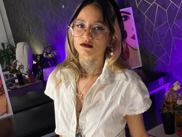 LiiaSmith, una mujer asiática con cabello rubio largo, que usa gafas y una blusa blanca, tiene un tatuaje floral grande en el cuello. Sus labios son de un rojo oscuro, y ella está mirando hacia adelante con una expresión sensual..