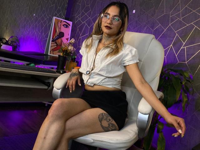 LiiaSmith está sentada em uma cadeira de escritório branca, com as pernas cruzadas, exibindo uma tatuagem na coxa. Ela veste uma camisa branca abotoada, uma saia preta, usa óculos e tem um olhar sexy e confiante. Iluminação em tons de roxo e preto.