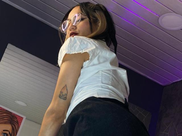 Liia Smith, con lunghi capelli neri, che indossa occhiali e una maglietta bianca a maniche corte, una gonna nera, un tatuaggio sul braccio sinistro, che sta guardando. in piedi, con il sedere visibile attraverso la gonna stretta, luci viola al soffitto.