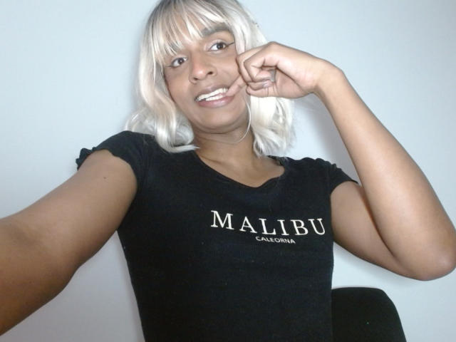 Fotografie von IdiiaLatina, einer schlanken, dunkelhäutigen Frau mit platinblondem Haar, die ein schwarzes T-Shirt mit der Aufschrift "MALIBU" trägt. Mit einem breiten Lächeln, das Kinn in der Hand ruhend, zeigte sie eine verführerische, kokette Pose..