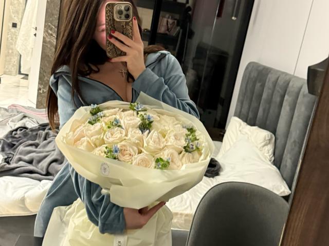 DenissaAnessa segurando um buquê de rosas cor de creme com pequenas flores verdes, vestindo uma camisa azul, tirando uma foto no espelho. Selfie com um celular dourado, em um quarto. Um buquê incrivelmente lindo..